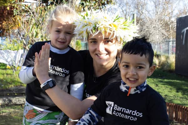 Amelia Kerr is wearing a Barnardos Aotearoa T-shirt and standing next to two smiling children.