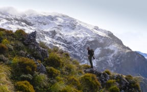 A wide shot of a man with a backpack and hiking poles standing on a rocky slope dotted with tussocks and low scrubby bushes. In the background is a snow-smattered rocky mountain.