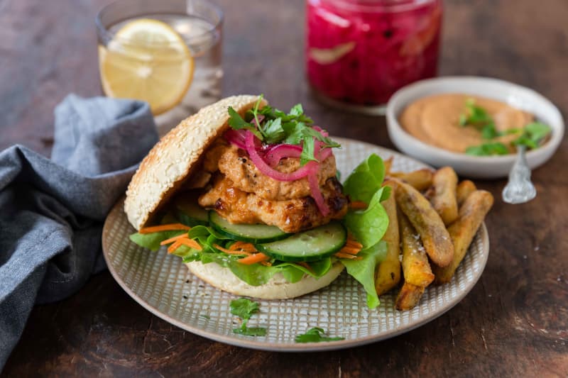 A Satay Chicken Burger - a split burger bun filled with lettuce, shredded carrot, cucumber, a satay chicken thigh and pickled red onions, with kumara chips on the side of the plate.
