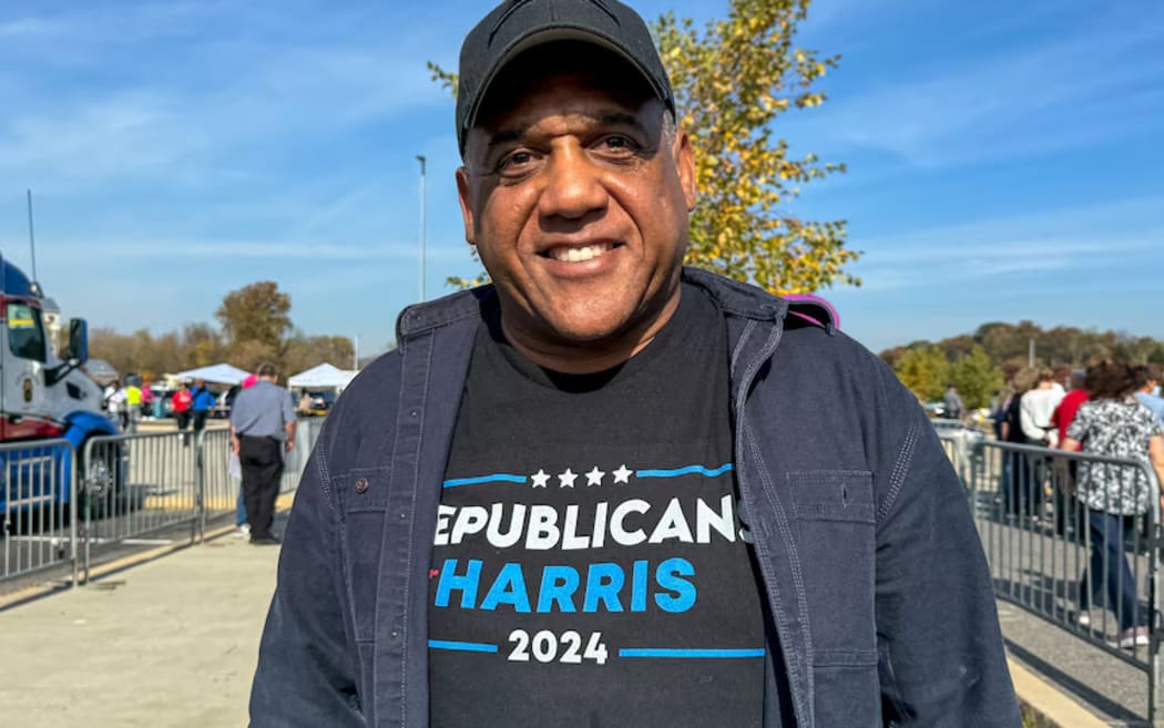 A man wears a 'Republicans for Harris' t-shirt while smiling and standing outside.