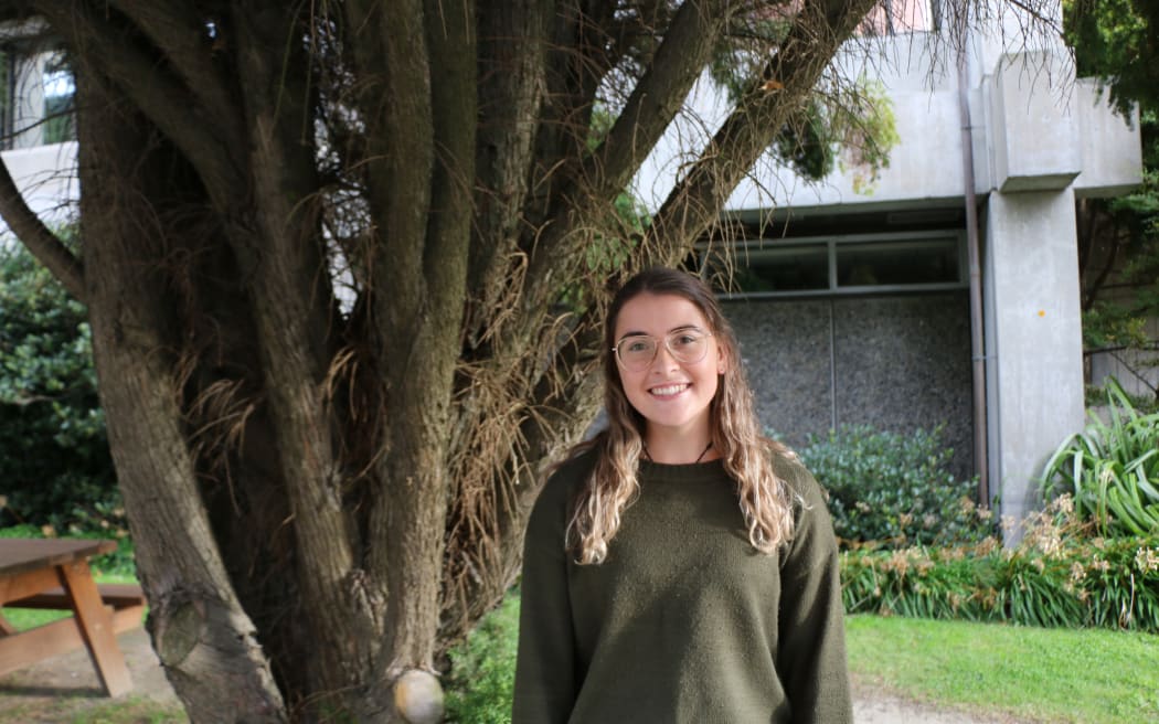 A midshot of a woman with long curly blond hair and wide-framed glasses in a khaki sweater standing in front of a tree and concrete building. She is looking at the camera and smiling.