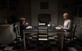 A mother and son sit at opposite ends of a dining table, in a dingy kitchen