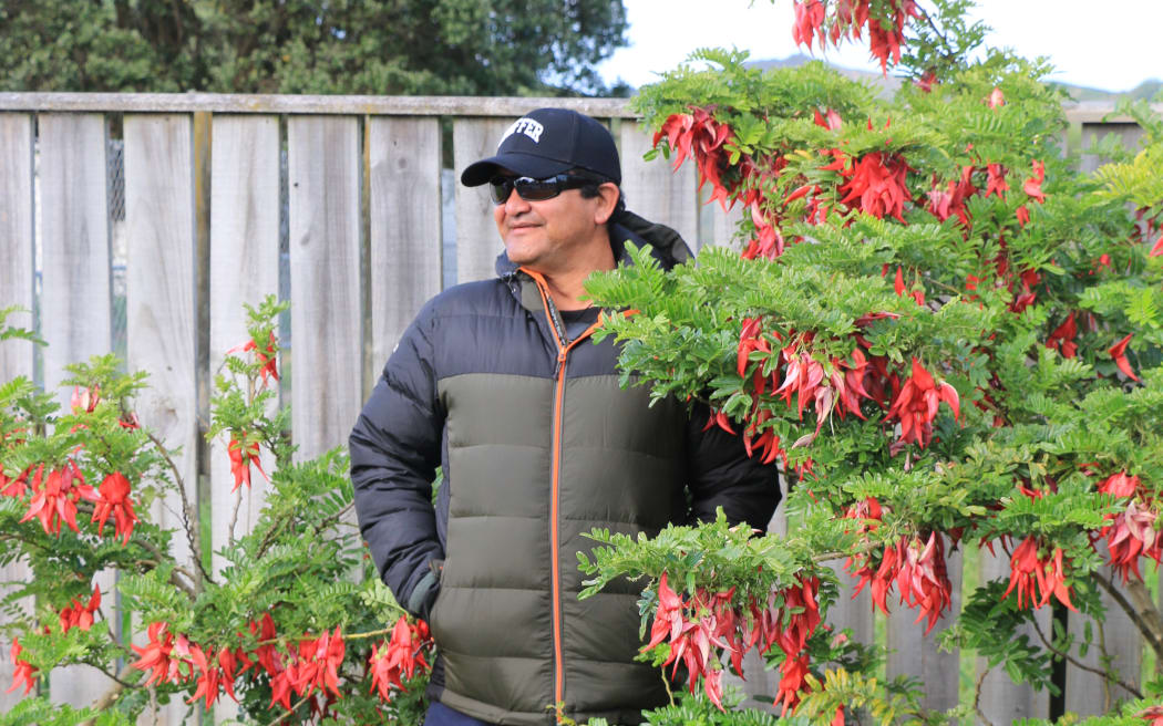 A man wearing a puffer jacket, cap and sunglasses stands next to a tall leafy bush with bright red flowers.