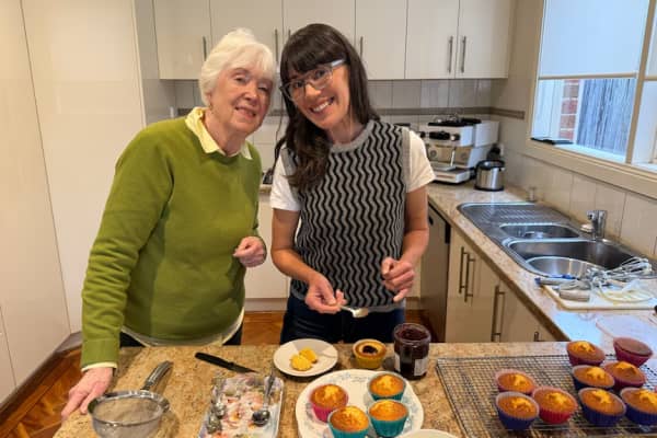 A white haired woman in a green sweaer and a long dark-haired woman in a grey vest stand together smiling over some fresh muffins.
