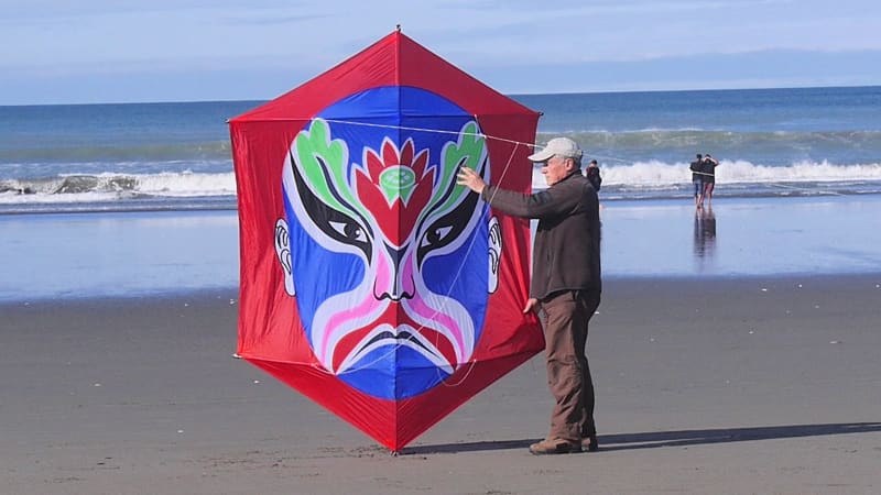 Jim Nicholls stands on the beach holding a giant red kite with a rokkaku design on it.