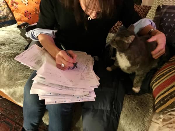 A person petting a cat while writing on a pile of paperwork in their lap.