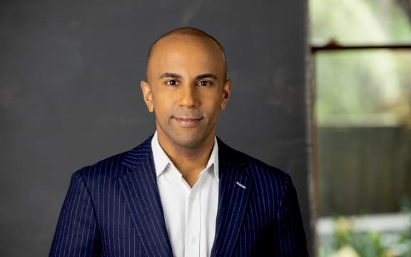 Jehan smiles at the camera. He is wearing a navy pinstripe suit and a white shirt. In the background in soft focus is the interior of a gently-lit house.