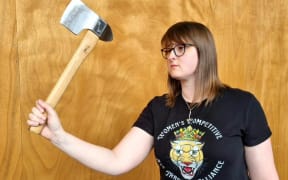 Britt holds an axe and looks poised to throw it. She is standing in front of a wooden wall and wearing a black tshirt.