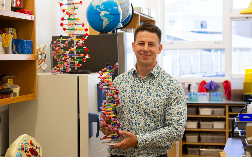 A midshot of Aidan holding a DNA structure model in front of a fridge and cupboards that are full of teaching equipment. Including two globes that sit high on a shelf.