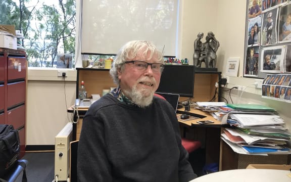 Warren Tate sitting at a desk in his office with photos on the wall and files on the top of shelves and tables behind him.