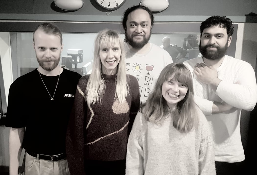 Five band members standing in the RNZ recording studio against the control room window