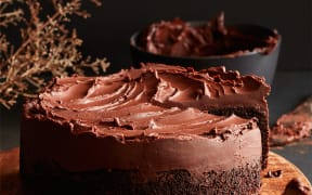 Image of a round chocolate cake with a thick head of chocolate ganache icing. The cake is on a wooden board in the foreground, with a bowl of ganache icing lurking at the back. A slice of cake is visible in the front righthand corner.
