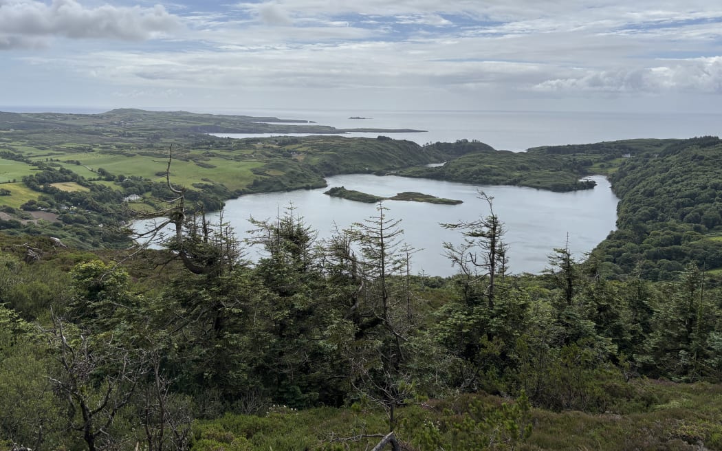 A view of the loch from above, looking out to the Atlantic coast. Three are two small islands in the centre of the loch, and the narrow path of the rapids is just visible connecting the inlet to the coast. It's surrounded by green fields and trees, beneath a partly cloudy sky.