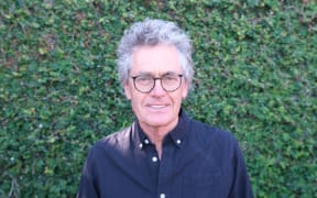 Mark smiles at the camera. He stands in front of a hedge wearing a black shirt and glasses.