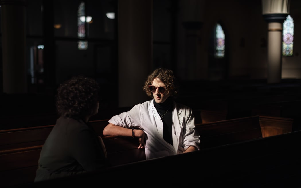 Presenter Ari Kerssens is seated in a church wearing a white shirt and glasses. He is surrounded by darkness.