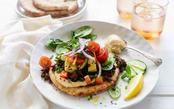 Spiced lamb mince with hummus and flatbreads on a round white plate garnished with herbs and a lemon wedge.