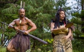 A still from Taki Rua Theatre: Breaking Barriers. Two actors stand amidst trees. On the left, a man wears feathers in his hair, a skirt, and holds a taiaha. On the right, a woman carries a patu, and wears a patterned skirt.