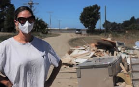 A woman wearing a white tshirt stands with her hands on her hips looking at the camera. She is wearing a KN95 face mask and black, cat-eye sunglasses. Behind her is a road covered in silt. The sky is blue.