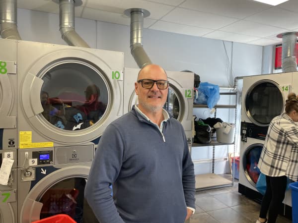 David Humphreys, owner of Auckland's Dayspring Laundromat. David is wearing glasses and a zip front sweatshirt, standing in a laundromat.