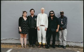 Five people stand against a grey wall. The man in the middle wears a white suit.