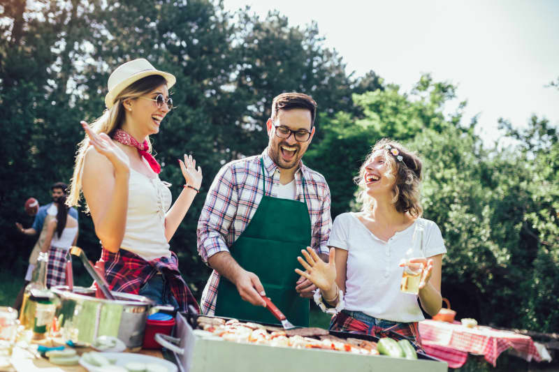 Group of people standing around grill, chatting, drinking and eating.
