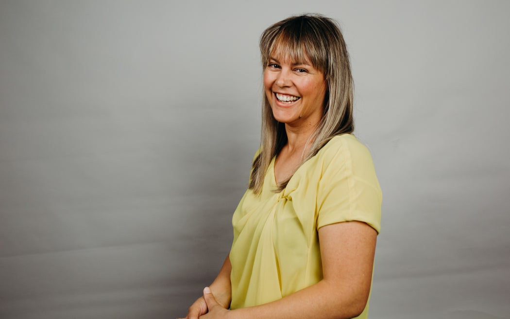 Dayna Vawdrey is standing in front of a grey background and smiling at the camera. She is weating a yellow blouse.