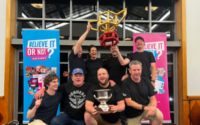 A pub quiz team dressed in black pose for a photo, some kneeling, some standing, holding aloft their trophy. They all smile.