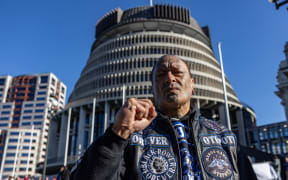 A Māori man with traditional face tattoos, wearing a black vest emblazoned with gang patches, stands in front of the Beehive in Wellington with his closed fist raised.