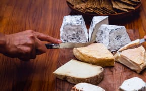 A close up of people cutting artisan cheeses with cheese knives. Several cheeses are placed on a wooden board, with a bowl of crackers in the background.