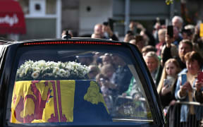 Members of the public pay their respects as they hearse carrying the coffin of Queen Elizabeth II, draped in the Royal Standard of Scotland, is driven through Ballater, on September 11, 2022. - Queen Elizabeth II's coffin will travel by road through Scottish towns and villages on Sunday as it begins its final journey from her beloved Scottish retreat of Balmoral. The Queen, who died on September 8, will be taken to the Palace of Holyroodhouse before lying at rest in St Giles' Cathedral, before travelling onwards to London for her funeral. (Photo by Paul ELLIS / AFP)