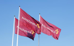 Three purple flags with the logo for Atrazeneca flying in the wind on top of the flagpoles against the blue sky.