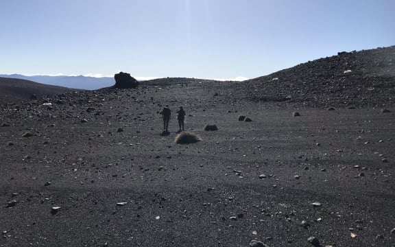Two people holding equipment stand in the mid ground. The scenery is dark gravel and rocks, it looks very much like a moonscape, with a smattering of plants. There's a clear blue sky in the background.