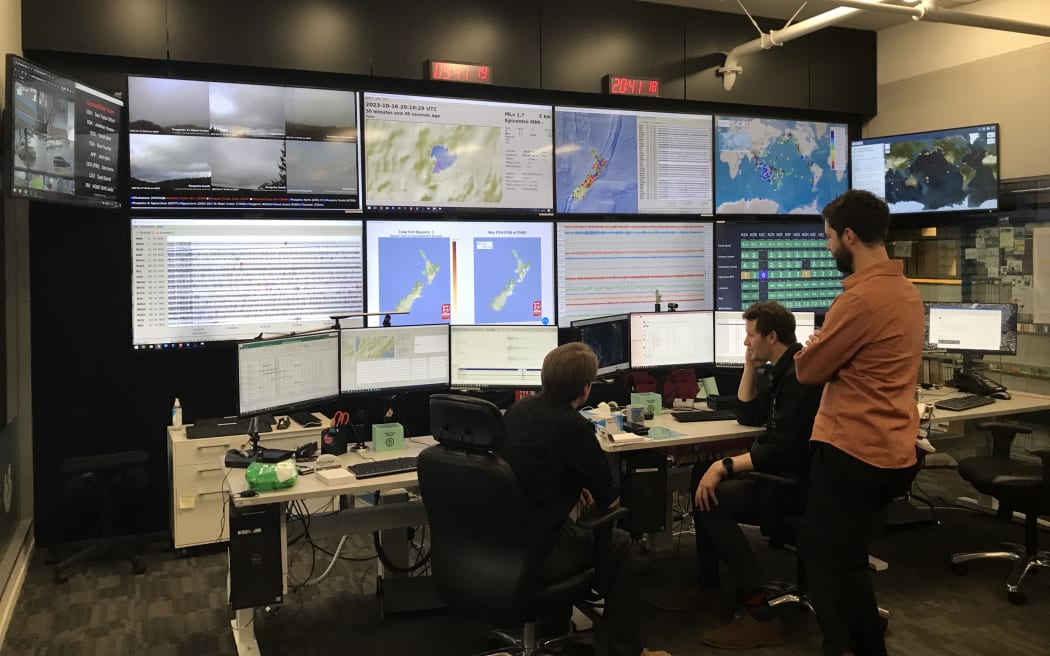 Three people look at an array of six computer monitors and 10 larger screens displaying maps and data.