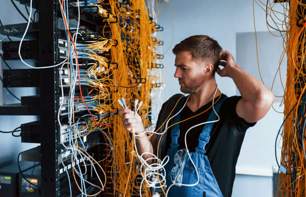 A man looks at a switchboard while holding several loose cables.