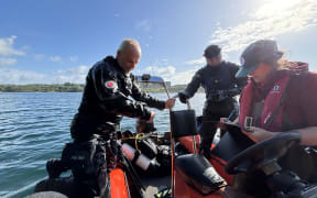 Three people on a small boat; two in diving suits and one writing on a clipboard. The boat is on calm water under a bright sky with scattered clouds.