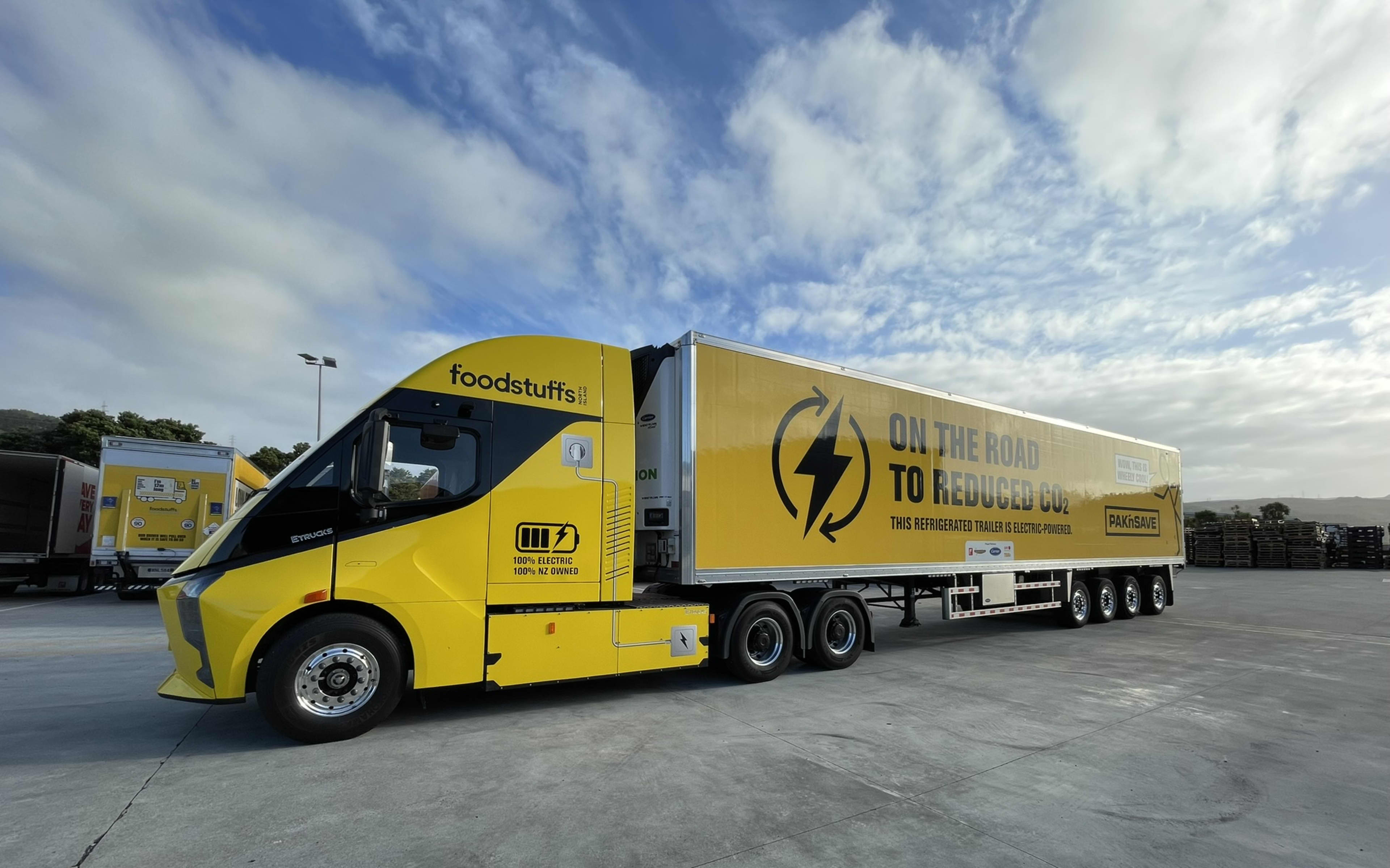 A bright yellow large electric truck and trailer unit with Foodstuffs branding is shown driving slowly out of a freight depot
