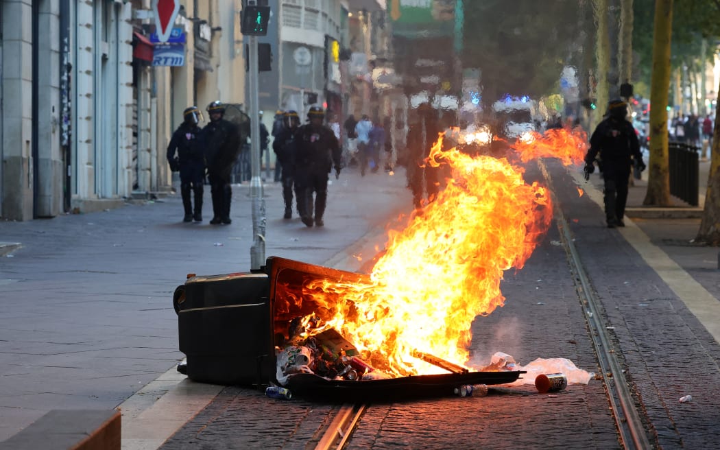 French riot police officers stand guard behind a burnt trash bin during a demonstration against police in Marseille, southern France on July 1, 2023, after a fourth consecutive night of rioting in France over the killing of a teenager by police. French police arrested 1311 people nationwide during a fourth consecutive night of rioting over the killing of a teenager by police, the interior ministry said on July 1, 2023. France had deployed 45,000 officers overnight backed by light armoured vehicles and crack police units to quell the violence over the death of 17-year-old Nahel, killed during a traffic stop in a Paris suburb on July 27, 2023. (Photo by CLEMENT MAHOUDEAU / AFP)