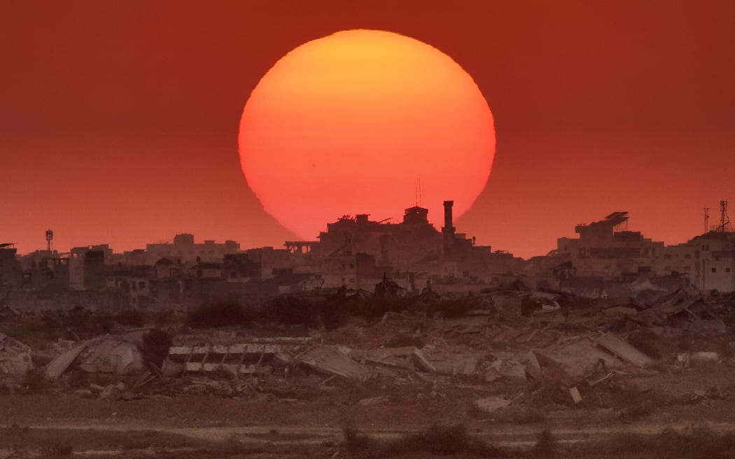 This picture taken from a position on the Israeli border with the Gaza Strip, shows the sun setting behind destroyed buildings in the besieged Palestinian territory on 19 August, 2025.