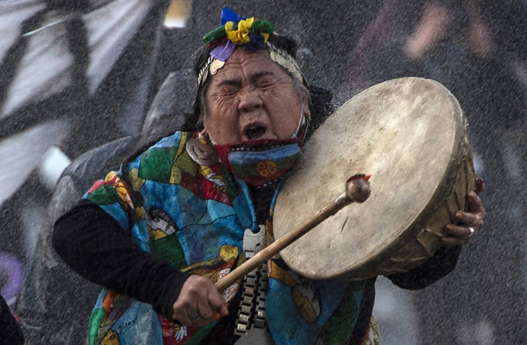 A Mapuche indigenous woman plays a drum as a riot police use water cannons to disperse demonstrators during a protest against the government's handling of the COVID-19 novel coronavirus pandemic, in Santiago.