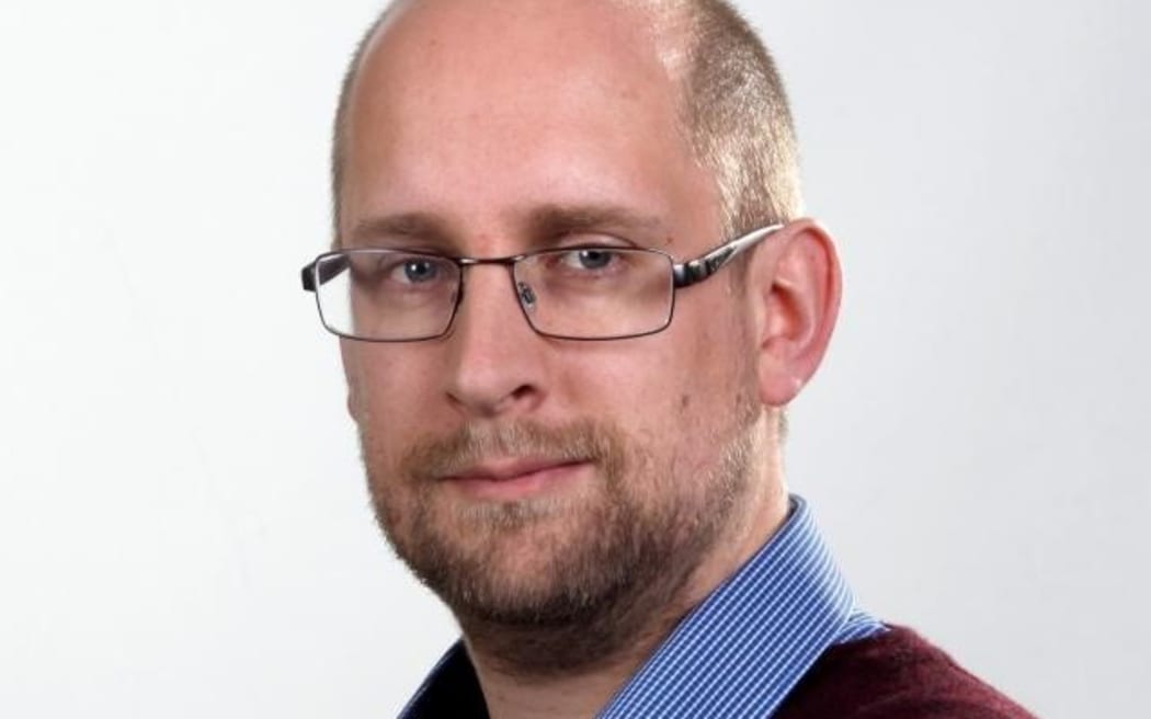 Dr Nic Rawlence looks intently at the camera. He is standing side-on to the camera, visible from the shoulders up, wearing a collared shirt and glasses.