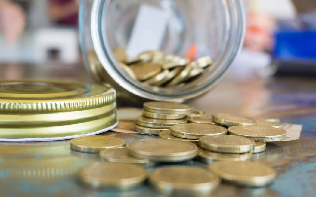 open tipped over savings jar with New Zealand currency coins spilling on to rustic wooden on wooden table closeup.