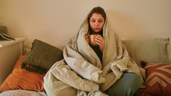 Woman looking tired and sick, wrapped up in blanket on couch, holding a mug.