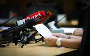 A submitter reads their written submission to a select committee and a lot of media microphones. Tissues on the table indicate the topic is sensitive.