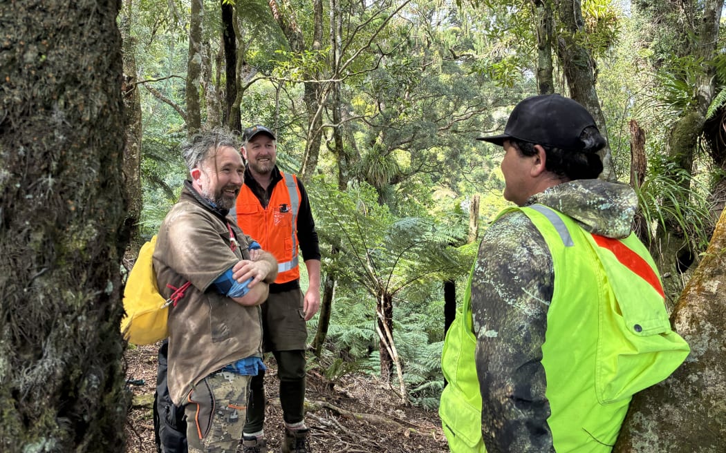 Three men stand in a forest, two facing the camera smiling, the third with his back to the camera. Two are wearing baseball caps and high-vis vests (one yellow, one orange), the third is wearing a brown jumper, with his arms crossed, and with a small yellow backpack on.