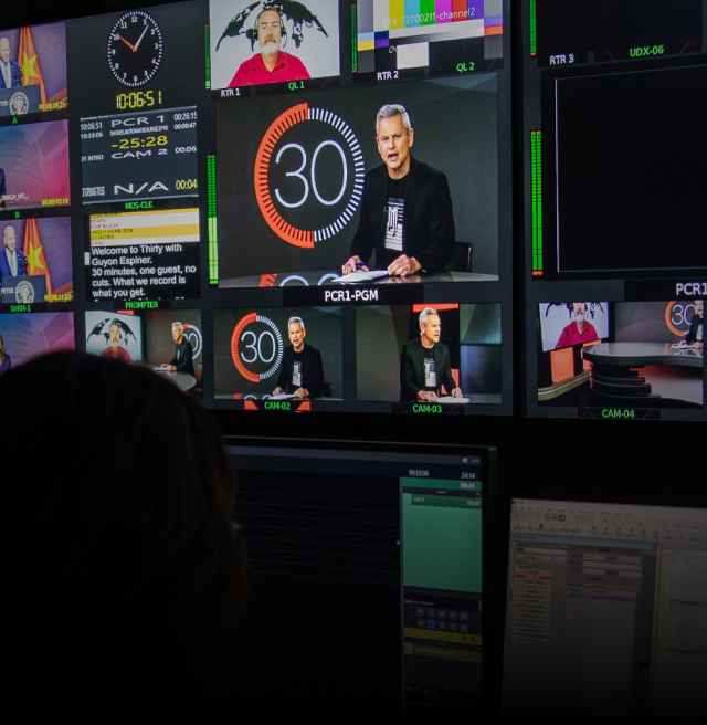 A darkened TV control room with a wall of monitors showing various inputs and camera views, in the centre is a minor with a man speaking in front of a large 30 logo.