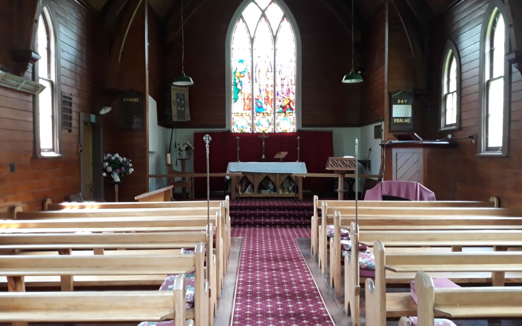 Interior of St Michael and All Angels Church, Porangahau. The carpeted aisle between the two blocks of wooden pews leads to the altar at the front of the church. A large stained glass window rises above the altar.