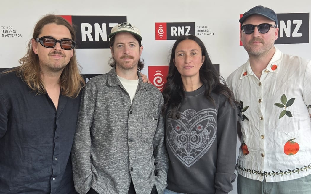 Three members of the band Leisure stand to have their picture taken with Kara Rickard in front of the RNZ wall.