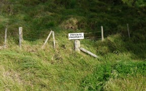 A sign that reads private property is hanging on a barbed wire fence. THe fence cuts across a grassy hill and is likely the boundry of a farm.