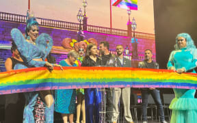 Drag Queens Miss Geena and Rita Menu hold the ceremonial rainbow ribbon, to be cut by Chlöe Swarbrick.