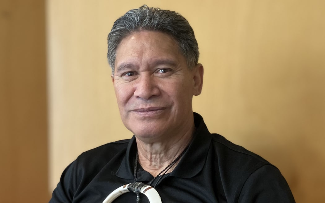 Blackie Tohiariki smiles at the camera and gives a thumbs up. He is wearing a black polo shirt and a large bone taonga around his neck.